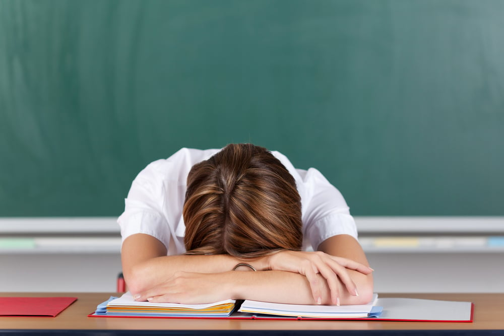 Teacher suffering from acute stress resting her head on her arms at her desk in front of the blackboard as she seeks to gather herself together